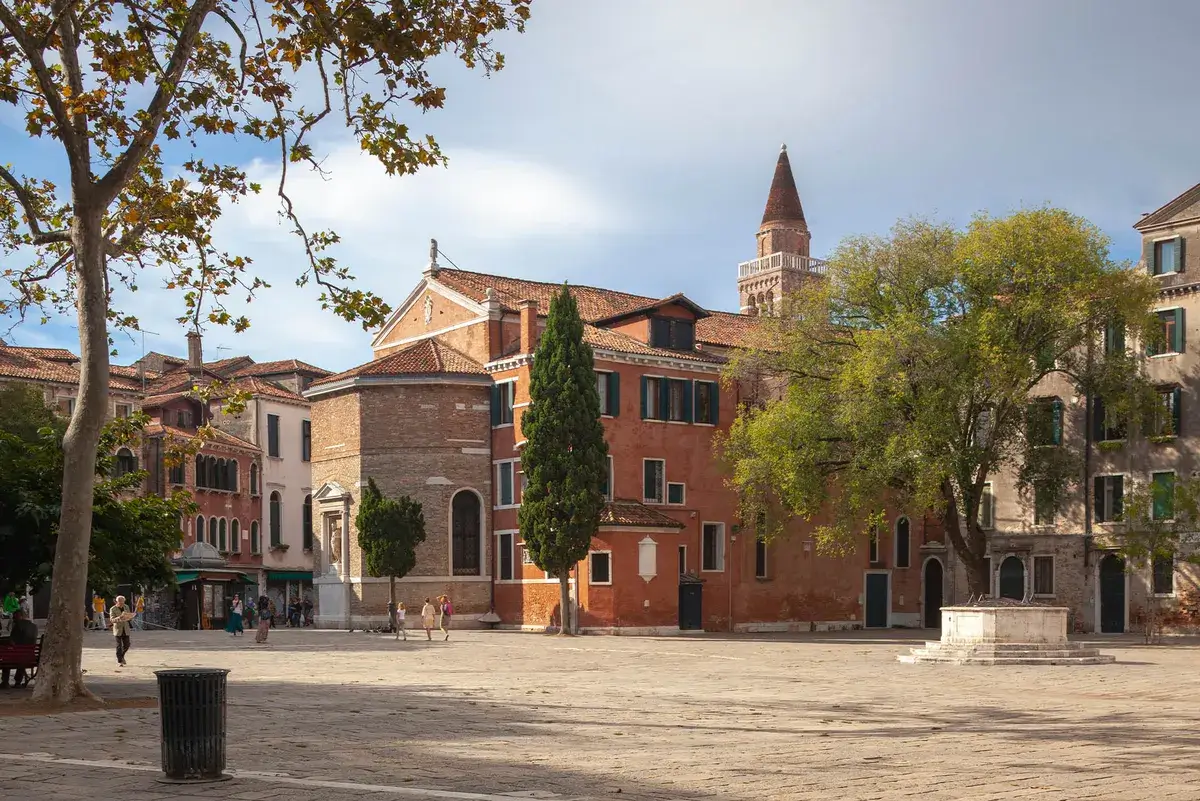 Piazza soleggiata con edifici storici, alberi, torre; alcune persone passeggiano nella piazza.