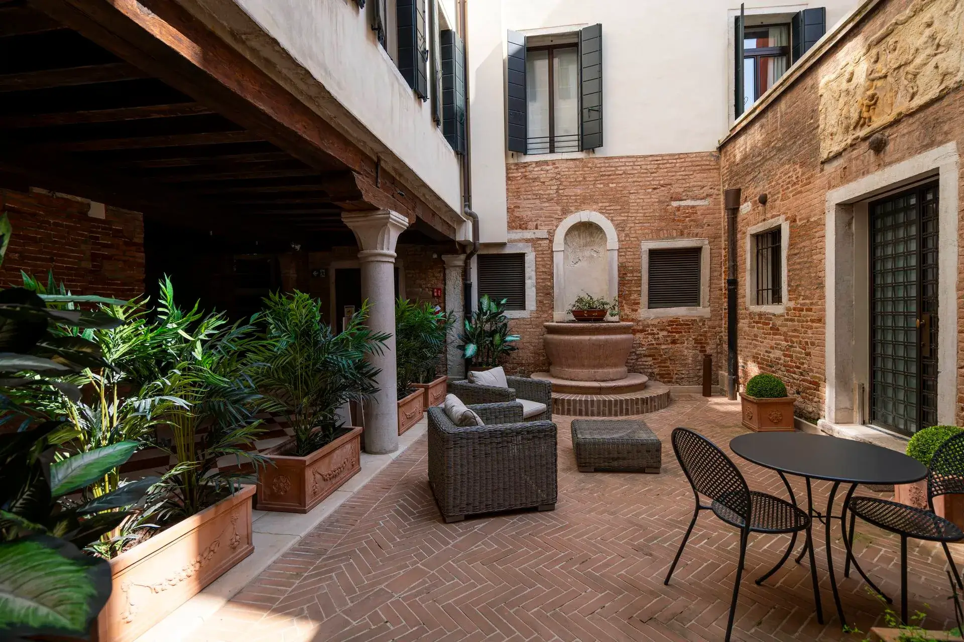 Cortile in mattoni: mobili da giardino, piante, fontana a muro; edifici con finestre e persiane.