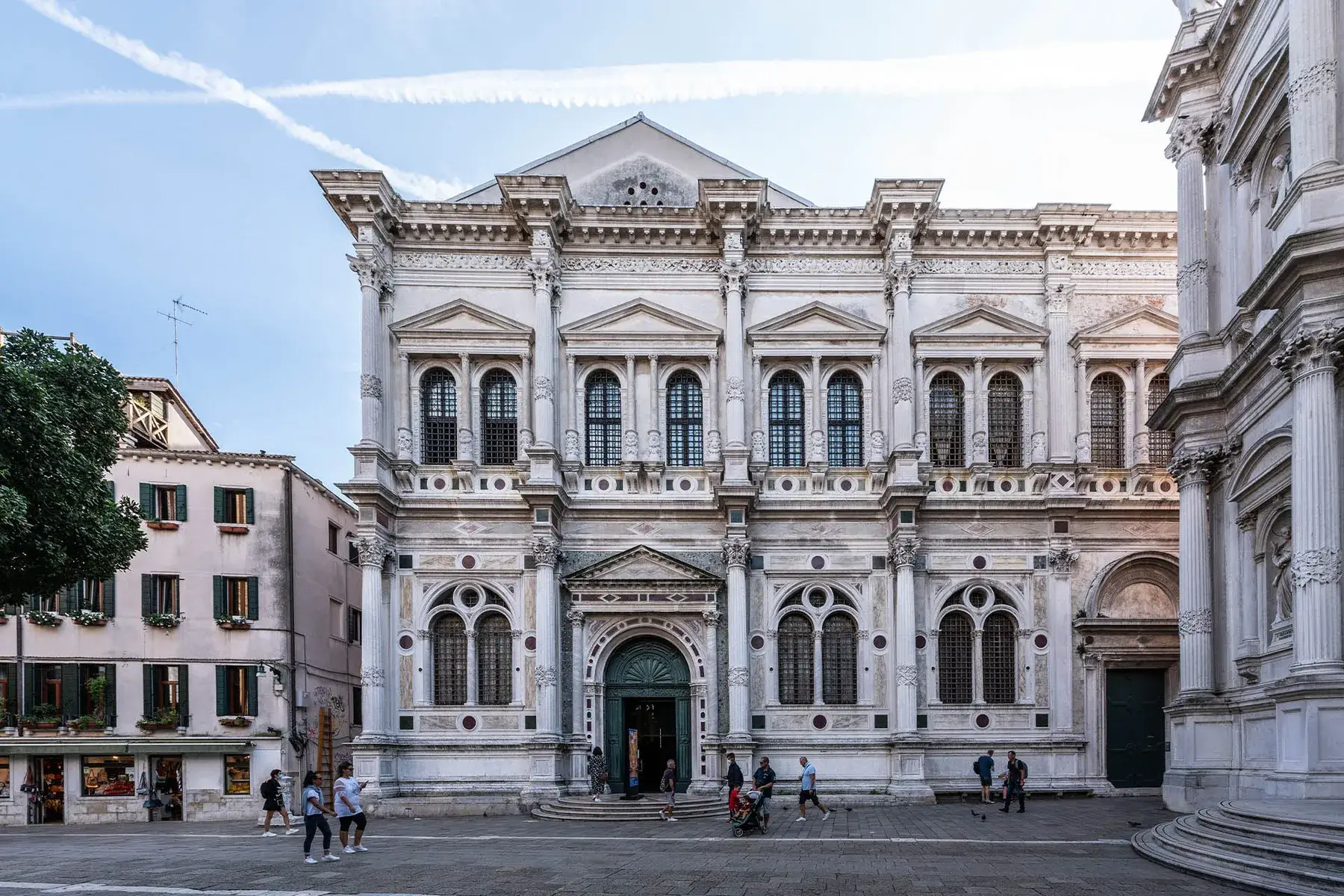 Edificio storico decorato con archi, colonne, persone in piazza, cielo azzurro.