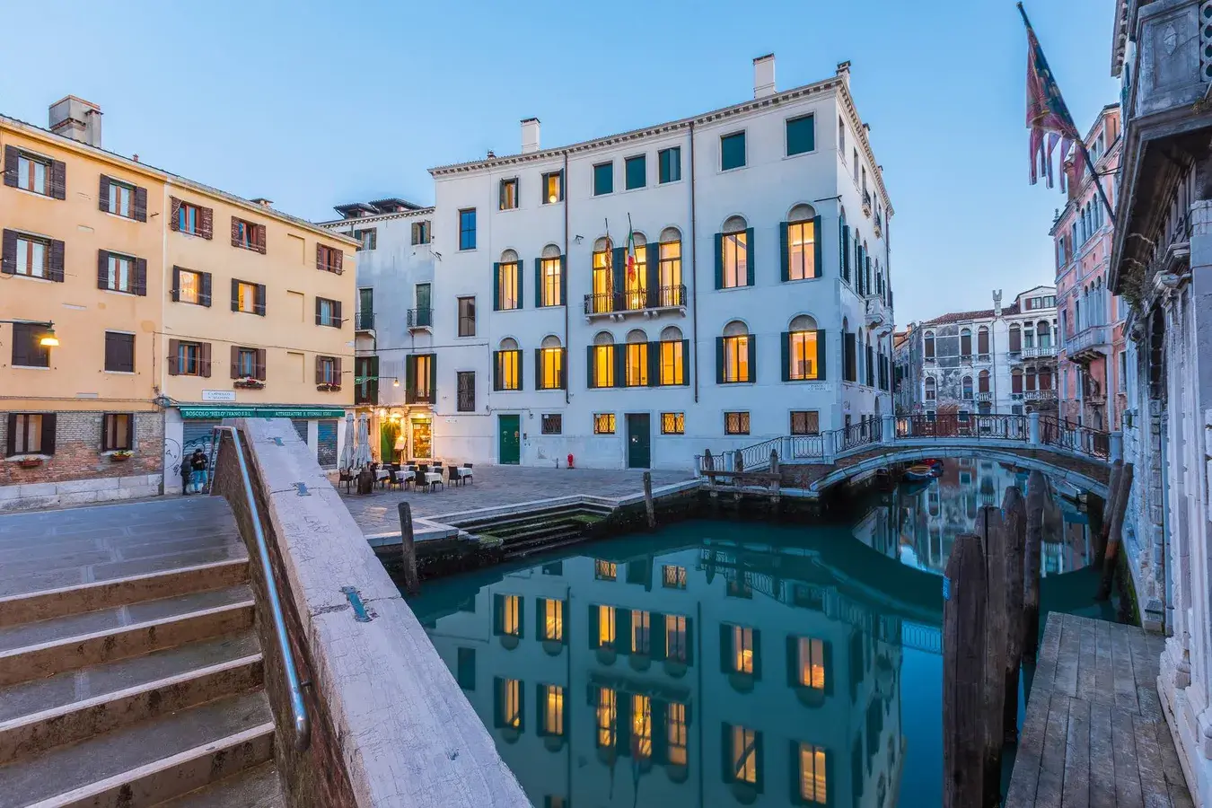 Canale di Venezia con edifici storici, riflessi, piccolo ponte; persone presso un bar sul lungomare.