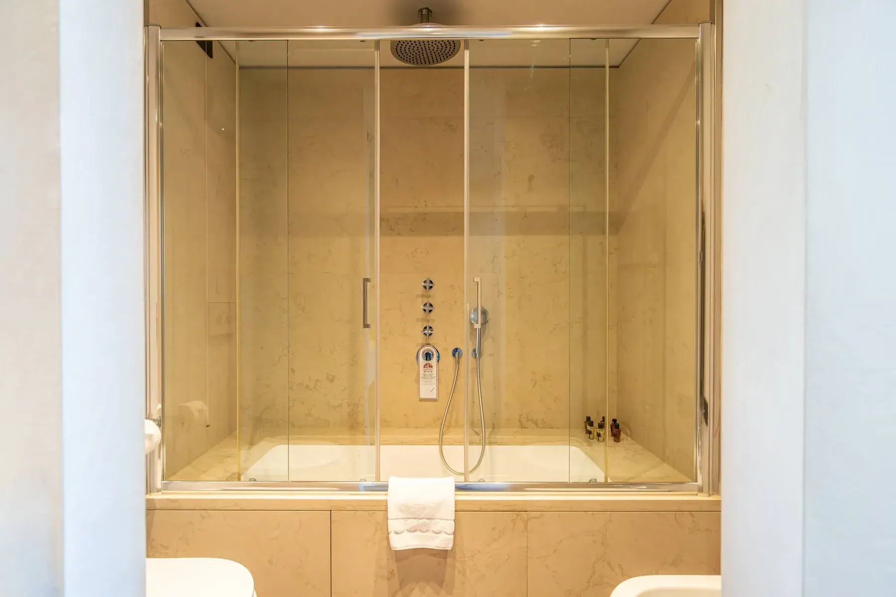 Bagno moderno: doccia in vetro, vasca, pareti in marmo beige, asciugamano appoggiato sul bordo della vasca.