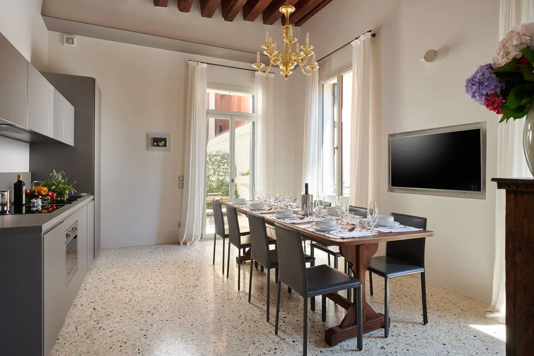 Sala da pranzo: lungo tavolo apparecchiato, cucina moderna, TV a parete e lampadario.