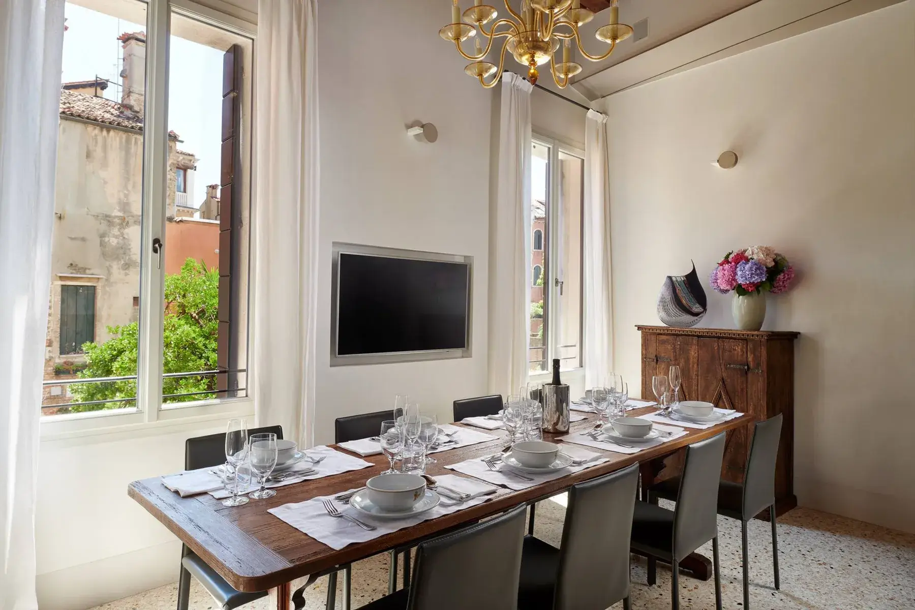 Sala da pranzo rustica con tavolo in legno, sedie nere, grandi finestre, lampadario e decorazioni.