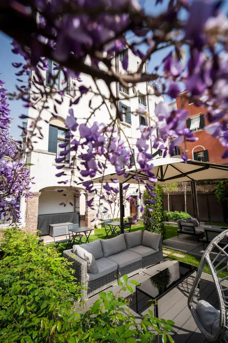 Patio accogliente: sedute in vimini, glicine viola e verde sotto un pergolato tra gli edifici.