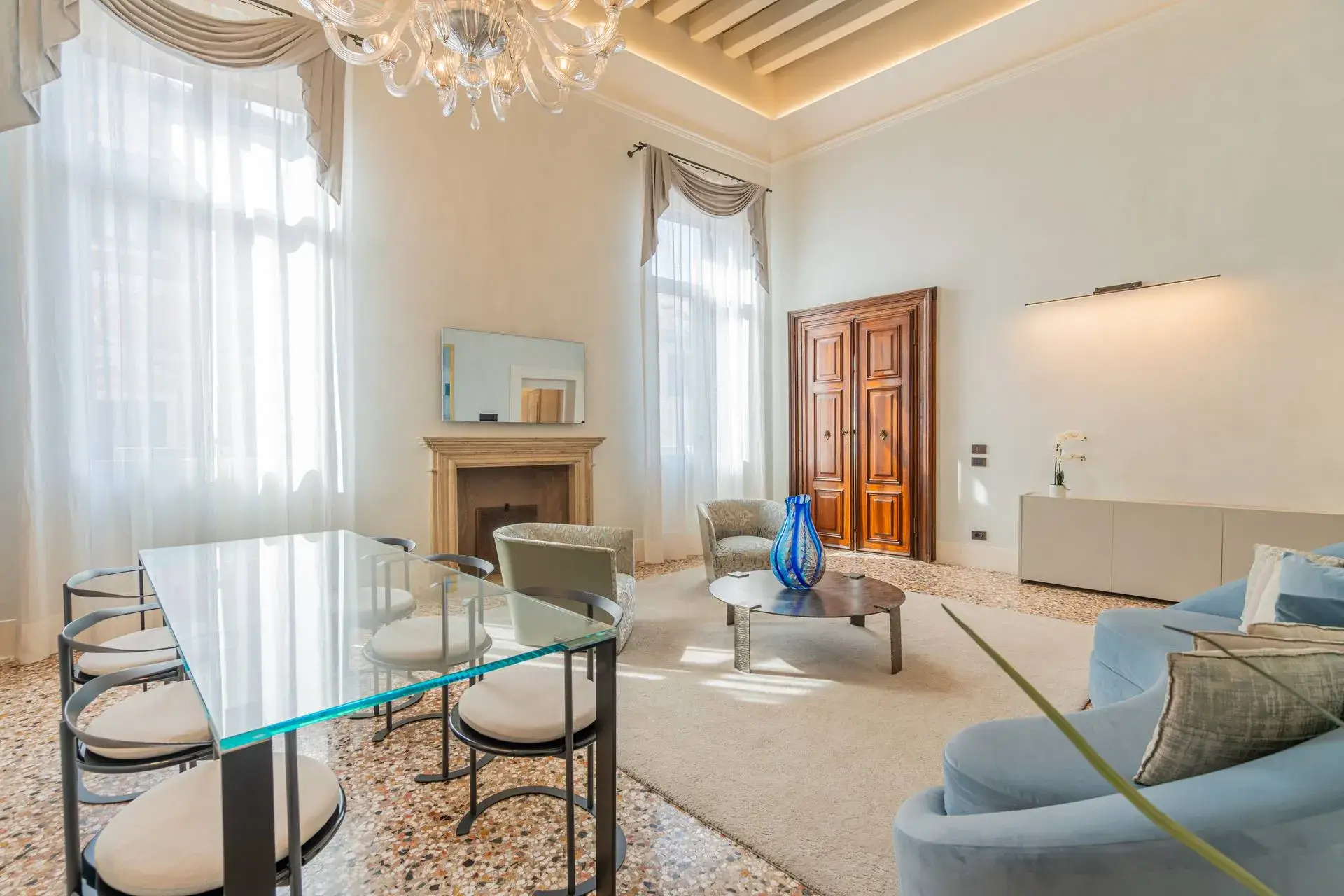 Chic living room: glass dining table, blue sofa, modern furniture and chandelier, high ceilings.