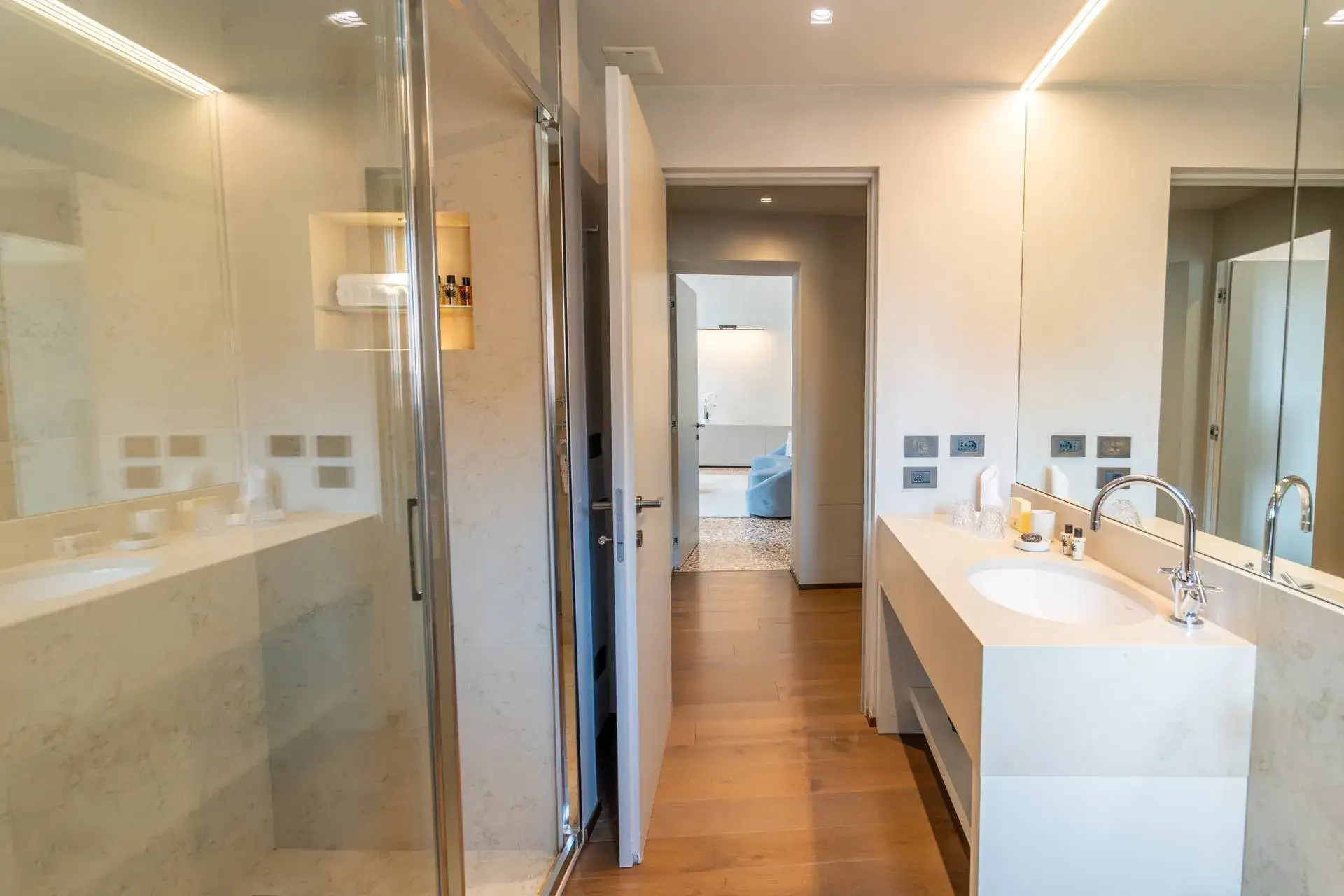 Modern bathroom: glass shower, large mirror, sink; leads to a bright room with wooden floors.