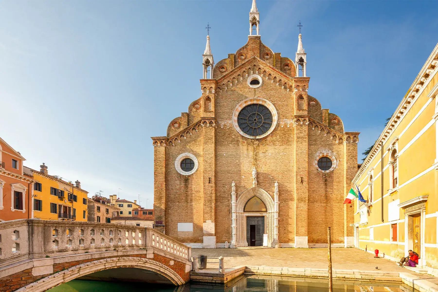 Una chiesa storica in mattoni, con rosoni, archi; accanto a un canale con gli edifici colorati di Venezia.