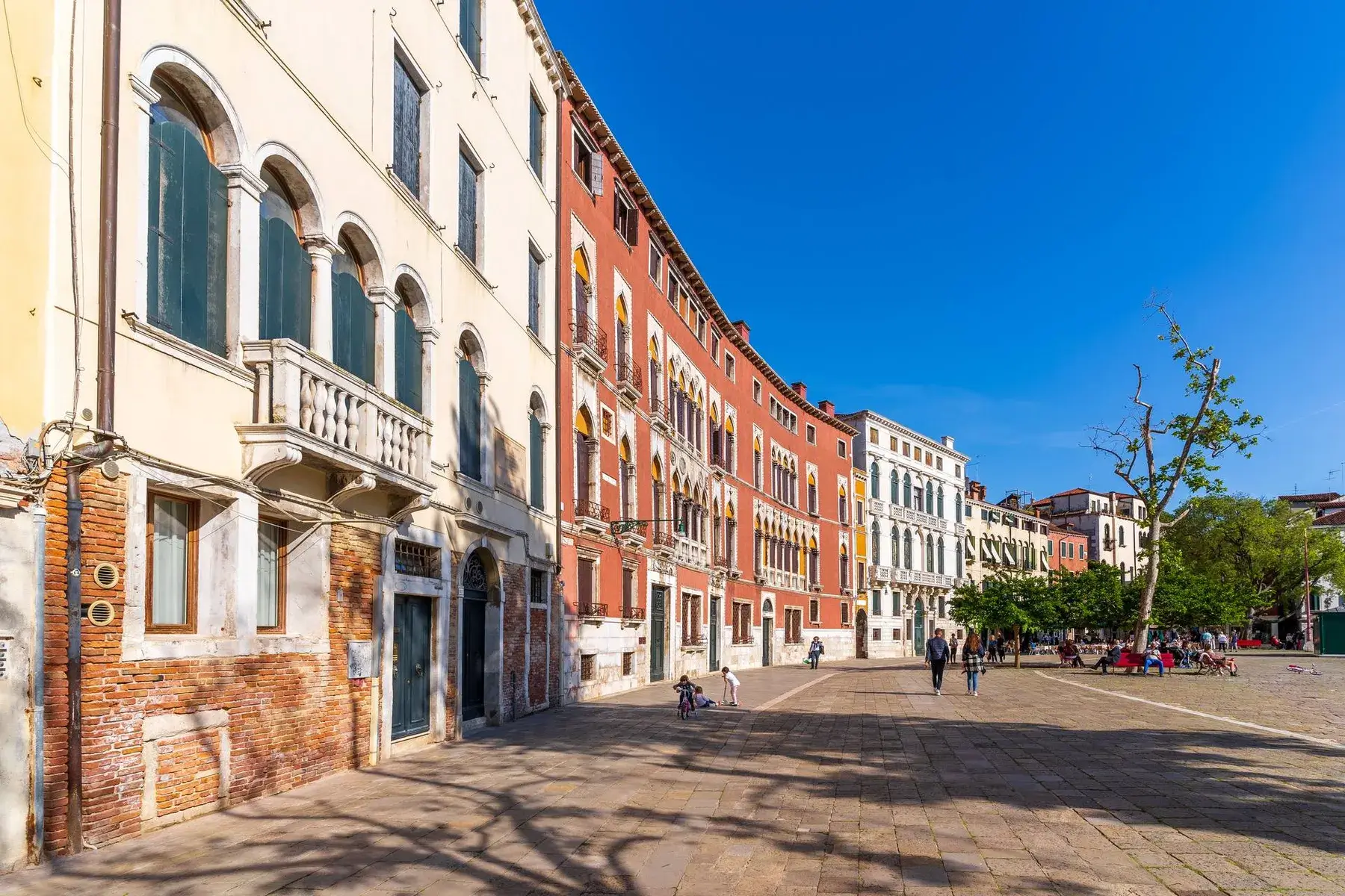 Edifici vivaci incorniciano una piazza soleggiata con persone, alberi e uno sfondo di cielo azzurro.