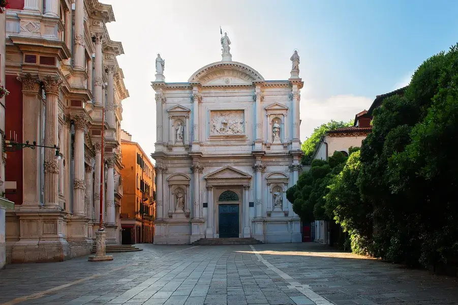 Chiesa in pietra bianca, facciata decorata, alberi ed edifici colorati sotto un cielo azzurro.