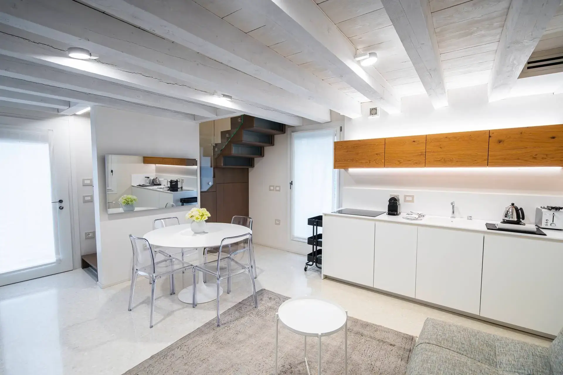 Cucina moderna con mobili bianchi, ripiani in legno e tavolo rotondo sotto il soffitto con travi a vista.