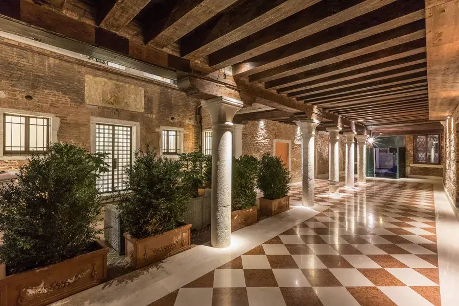 Corridoio con soffitto in legno, colonne in pietra, pavimento a scacchi; piante lungo le colonne illuminate.