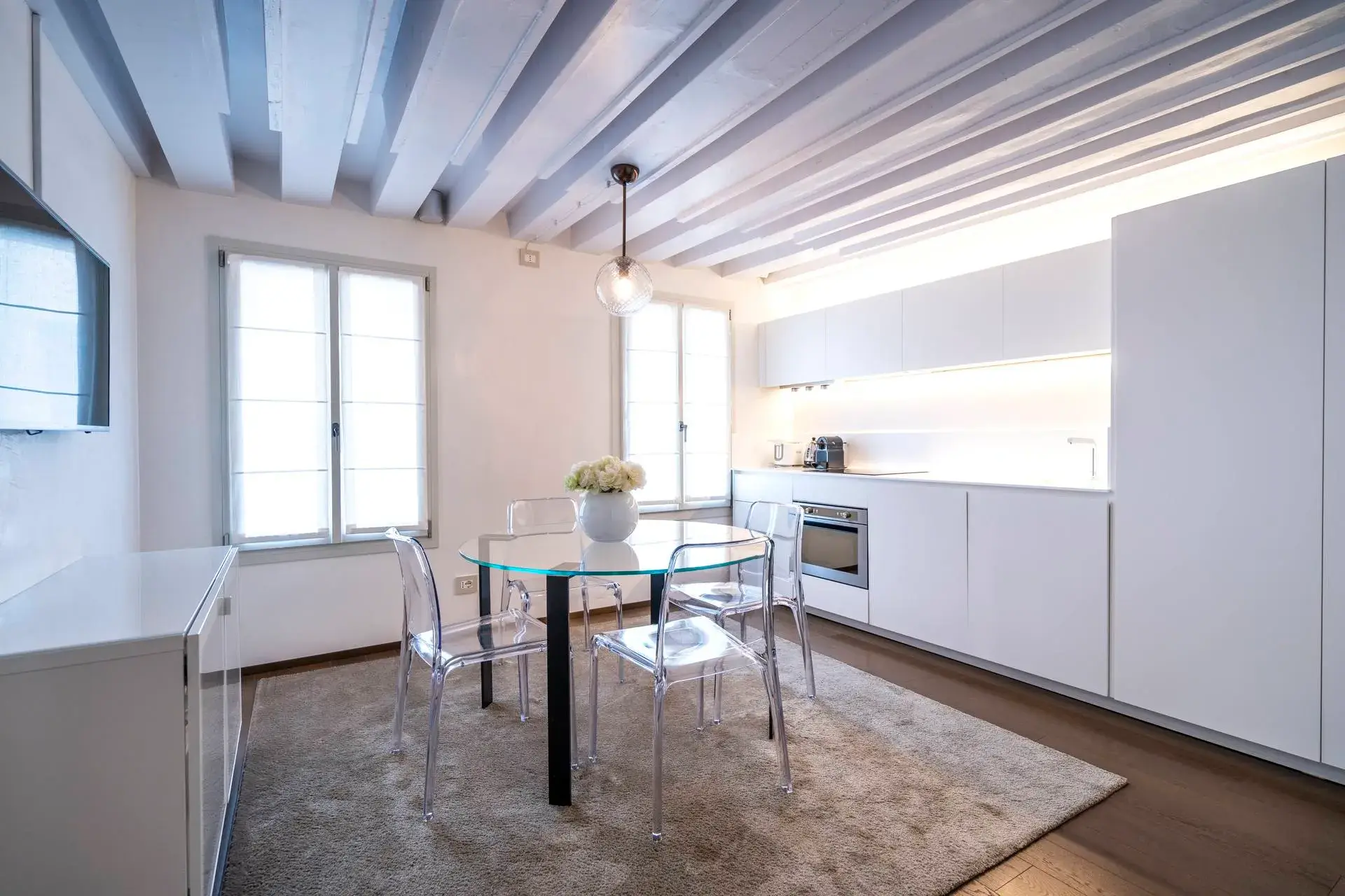 Minimalist kitchen: glass table, clear chairs, white cabinets, beamed ceiling.