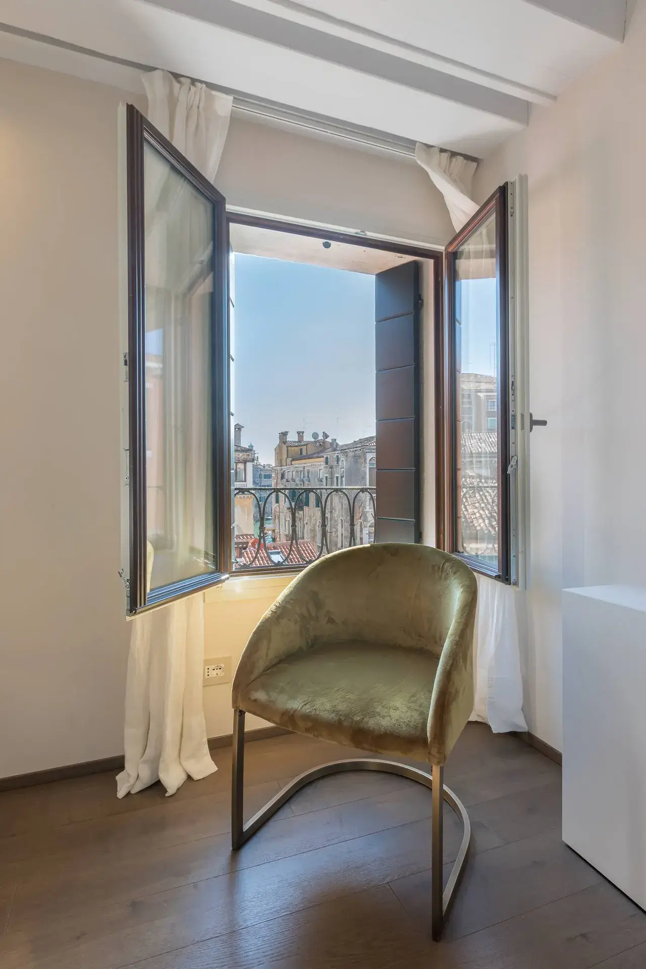 A velvet armchair by an open window with city views, framed in white curtains.