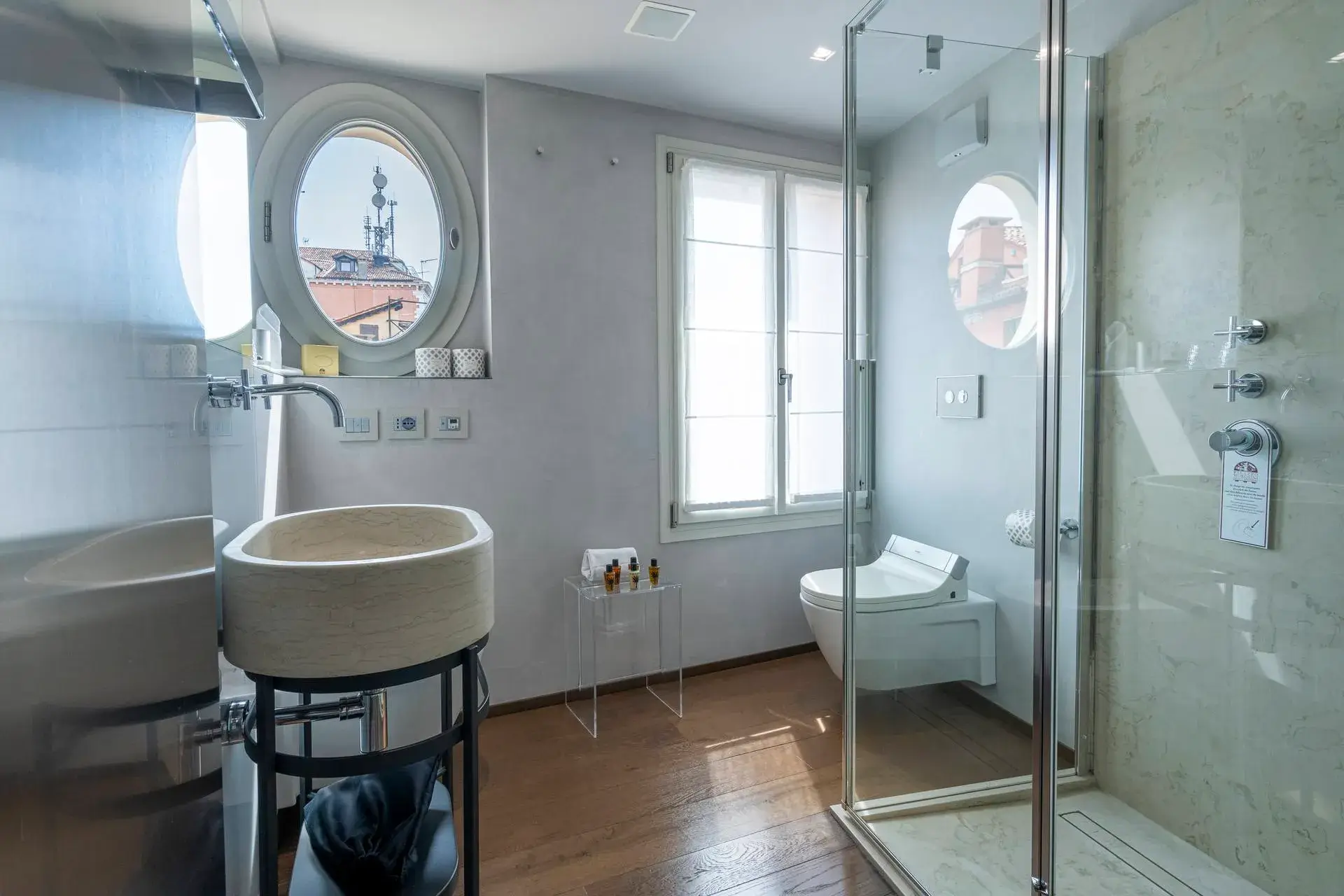 Modern bath: oval window, glass shower, wall sink, with city view.