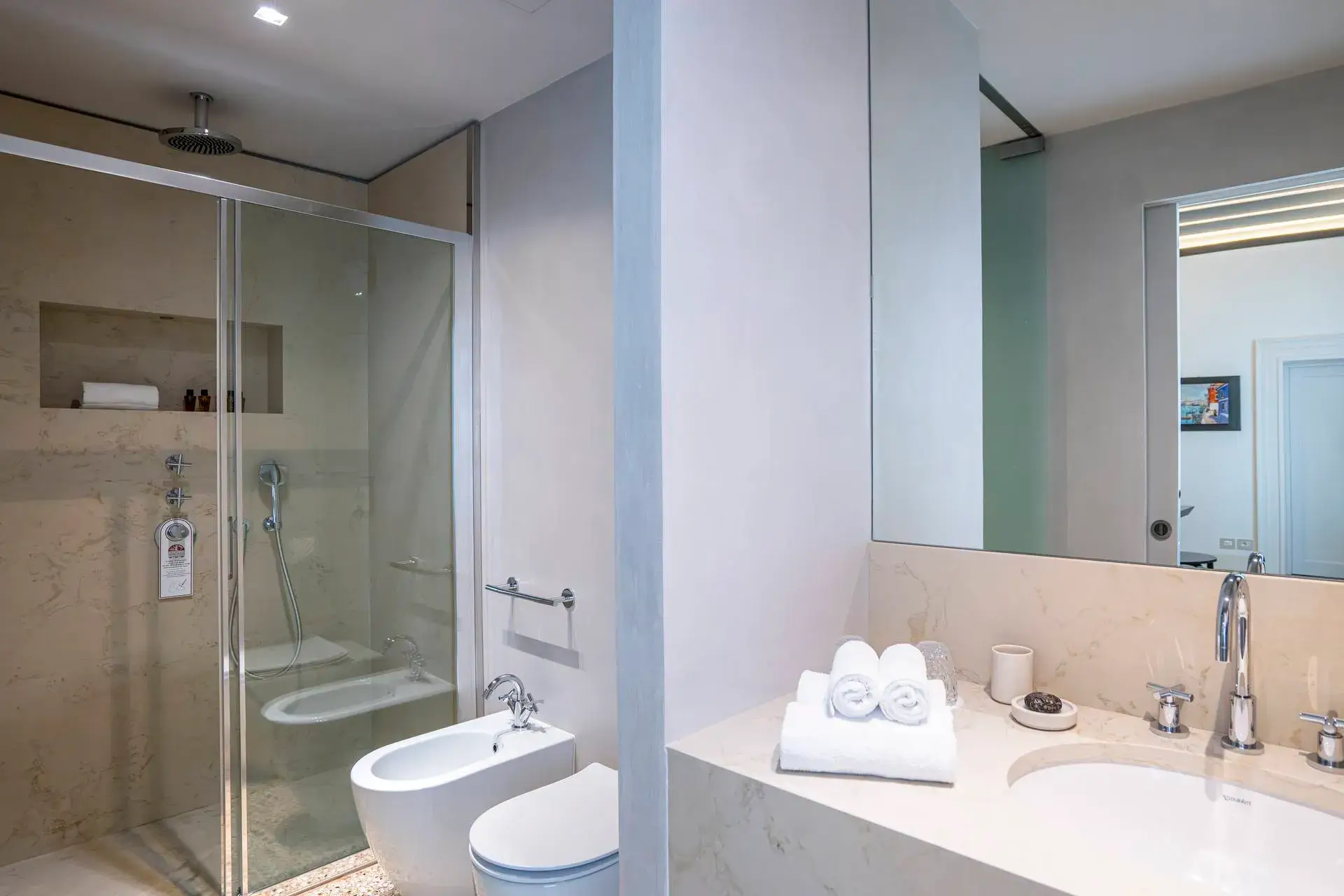 Modern bathroom: glass shower, toilet, bidet, sink with towels & toiletries on counter.