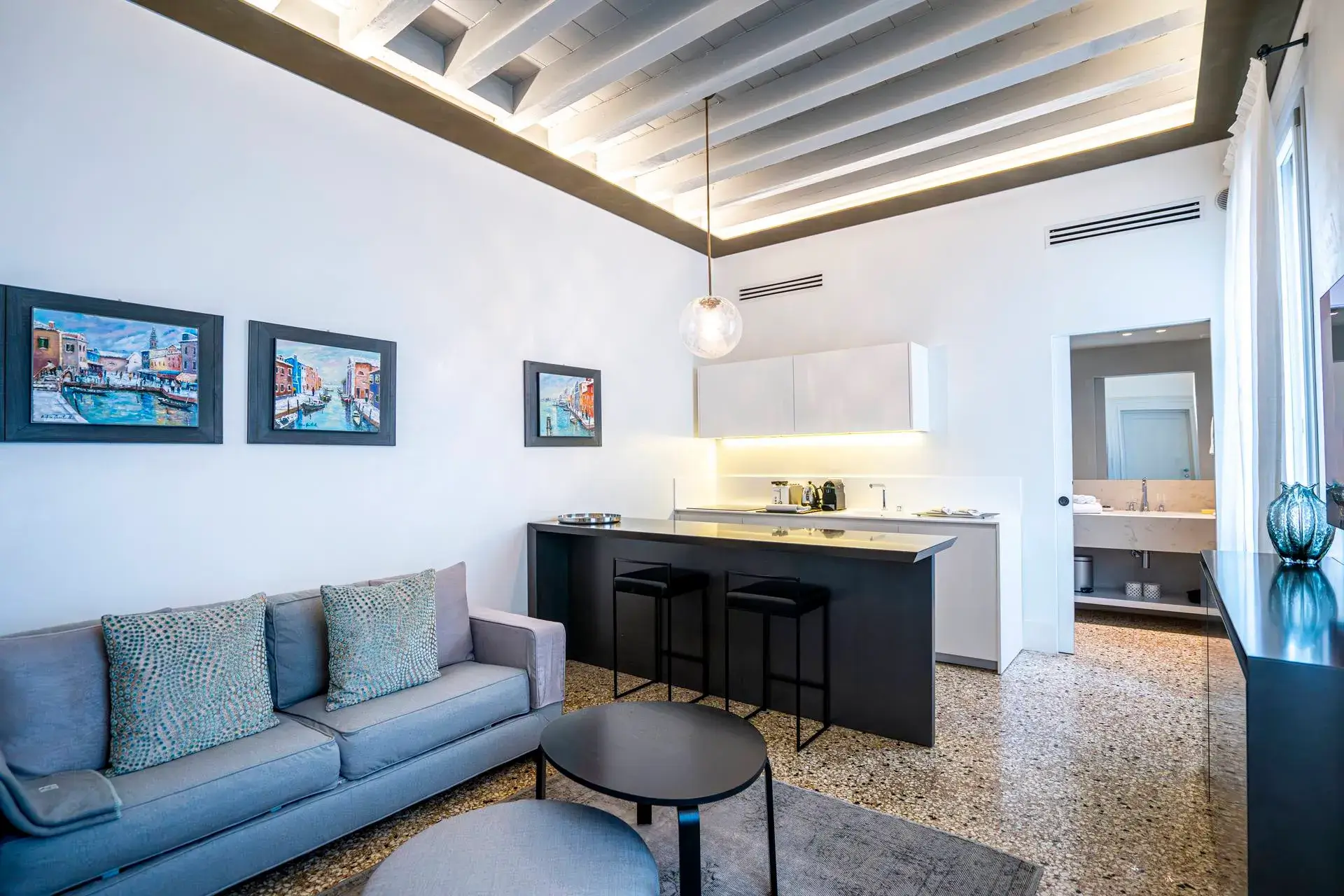 Modern living room: gray sofa, kitchen island, bar stools, colorful art on white walls.