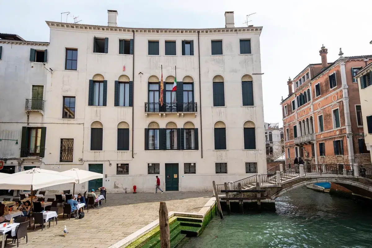 Una persona passeggia lungo un canale a Venezia, passando davanti a un edificio con finestre ad arco, una bandiera e un ponte.