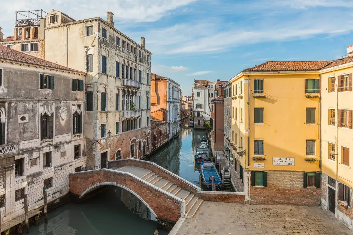 Una vista sul canale di Venezia con un ponte curvo, edifici colorati e piccole barche sotto il cielo azzurro.