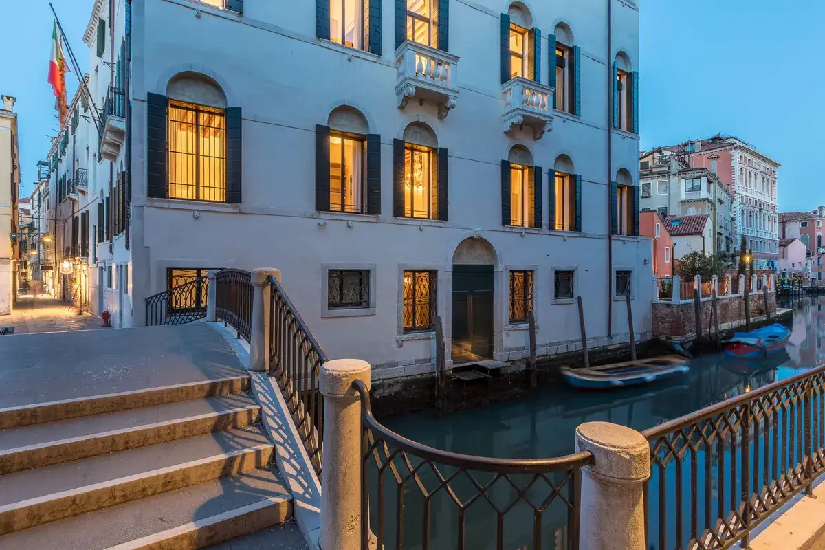 Canale di Venezia al tramonto: barche, finestre illuminate, edificio storico e ponte di pietra in vista.