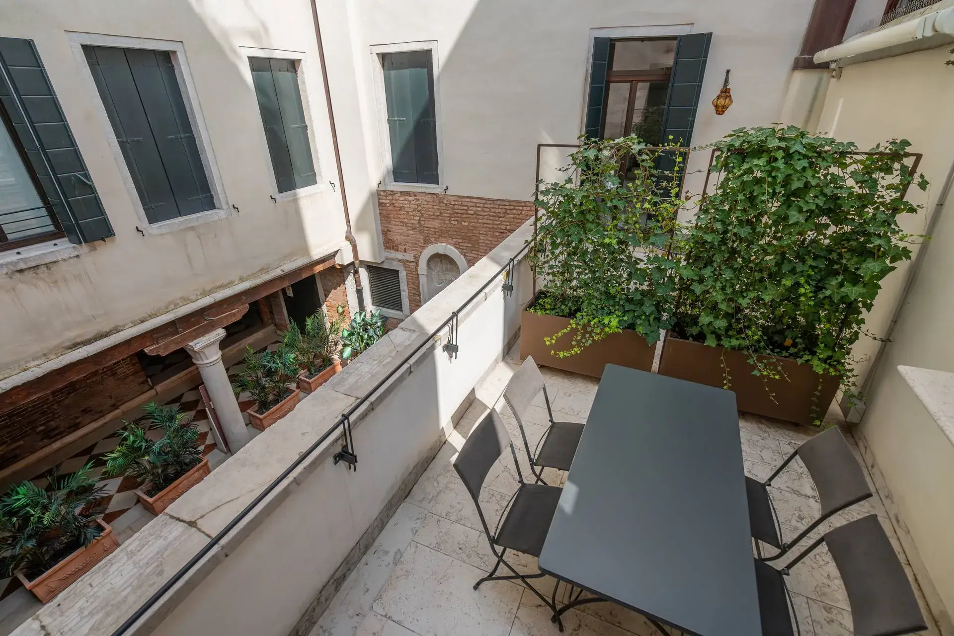 Terrazza soleggiata con tavolo, sedie e verde che si affaccia su un cortile ricco di piante.