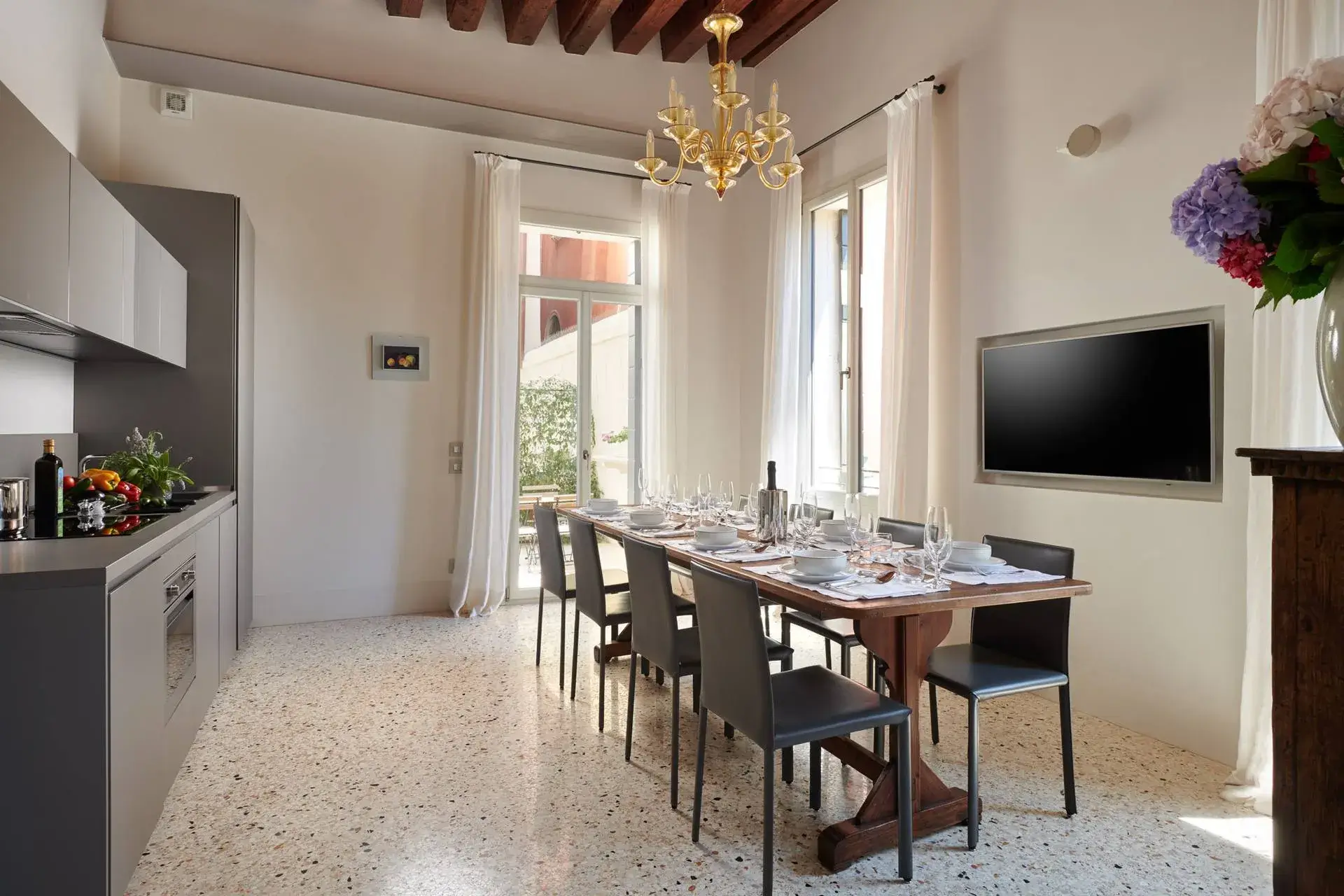 Sala da pranzo moderna: tavolo in legno, sedie nere, elegante lampadario, TV, pareti e tende bianche.